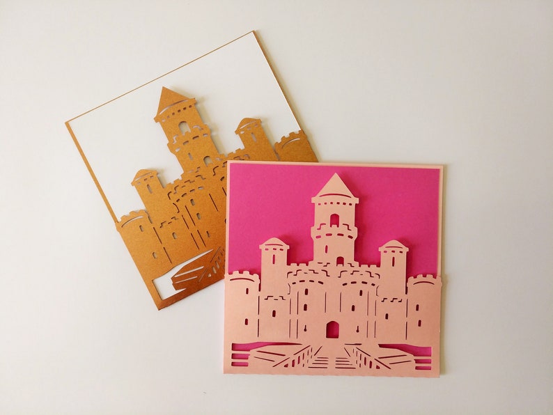 May include: Two paper cards with a castle design cut out. The top card is white with a gold castle design. The bottom card is pink with a pink castle design.