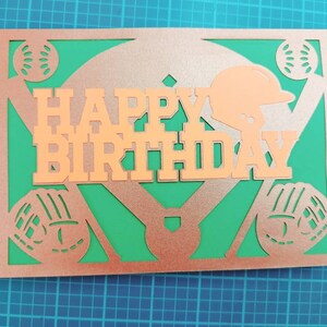May include: A copper-colored card with a baseball diamond design. The words "Happy Birthday" are cut out of the card and the background is green.