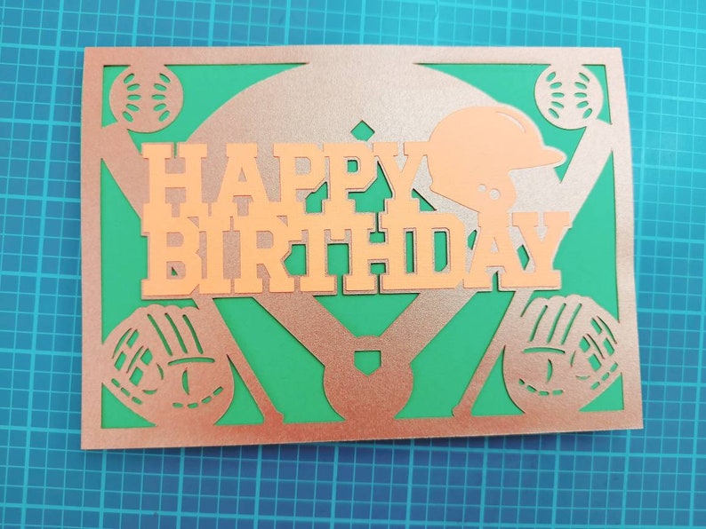 May include: A copper-colored card with a baseball diamond design. The words "Happy Birthday" are cut out of the card and the background is green.