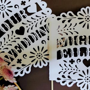 May include: White paper cutouts with intricate floral and heart designs. One cutout reads "OUR WEDDING" and the other reads "MIGUEL & EILEEN". Pink and white flowers are in the foreground. The cutouts are likely for a wedding.