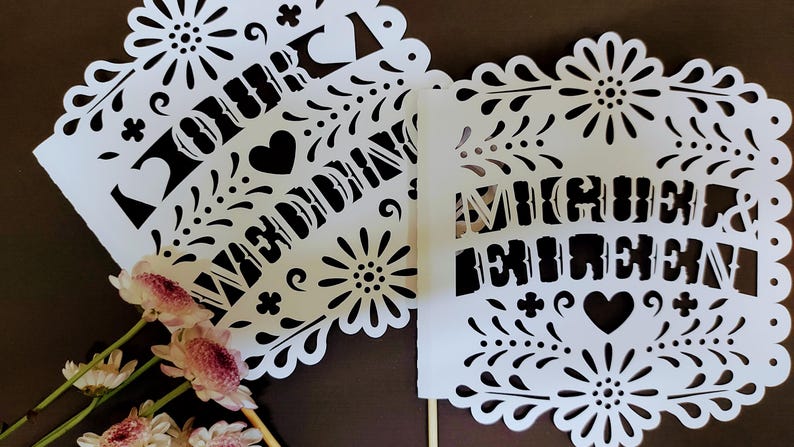 May include: White paper cutouts with intricate floral and heart designs. One cutout reads "OUR WEDDING" and the other reads "MIGUEL & EILEEN". Pink and white flowers are in the foreground. The cutouts are likely for a wedding.