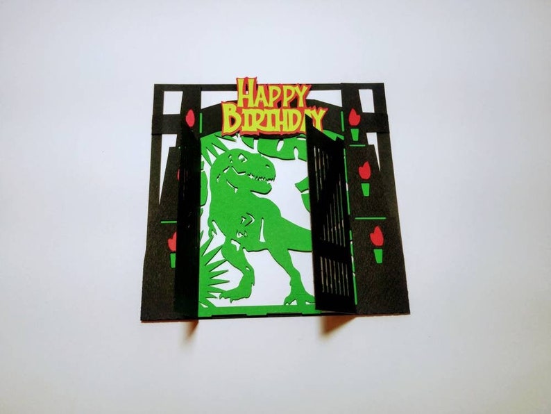 May include: A green dinosaur pops out of a black and red paper card with the words "Happy Birthday" on the top.