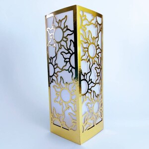 May include: Gold lantern with a cut-out design of suns. The lantern is made of paper and has a white interior.