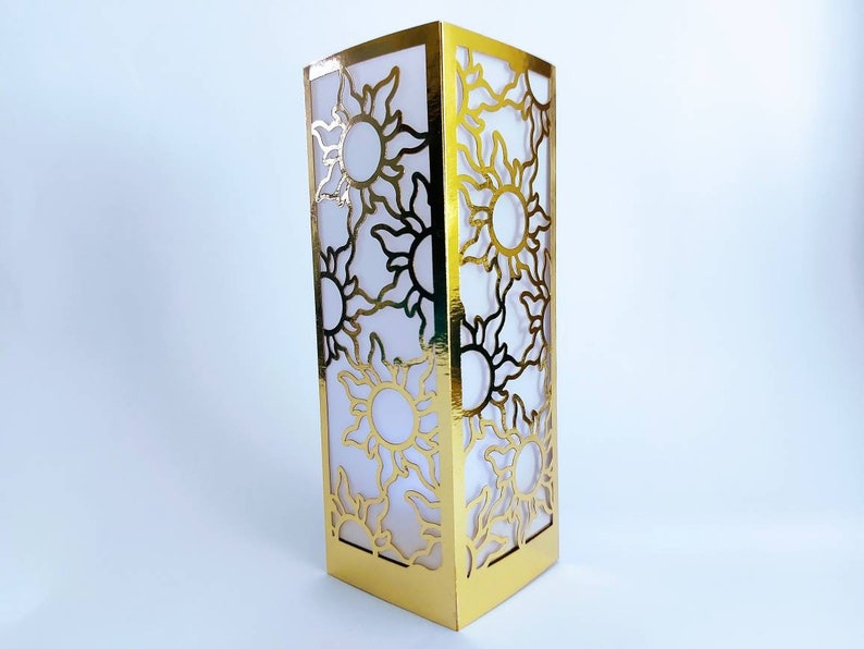 May include: Gold lantern with a cut-out design of suns. The lantern is made of paper and has a white interior.
