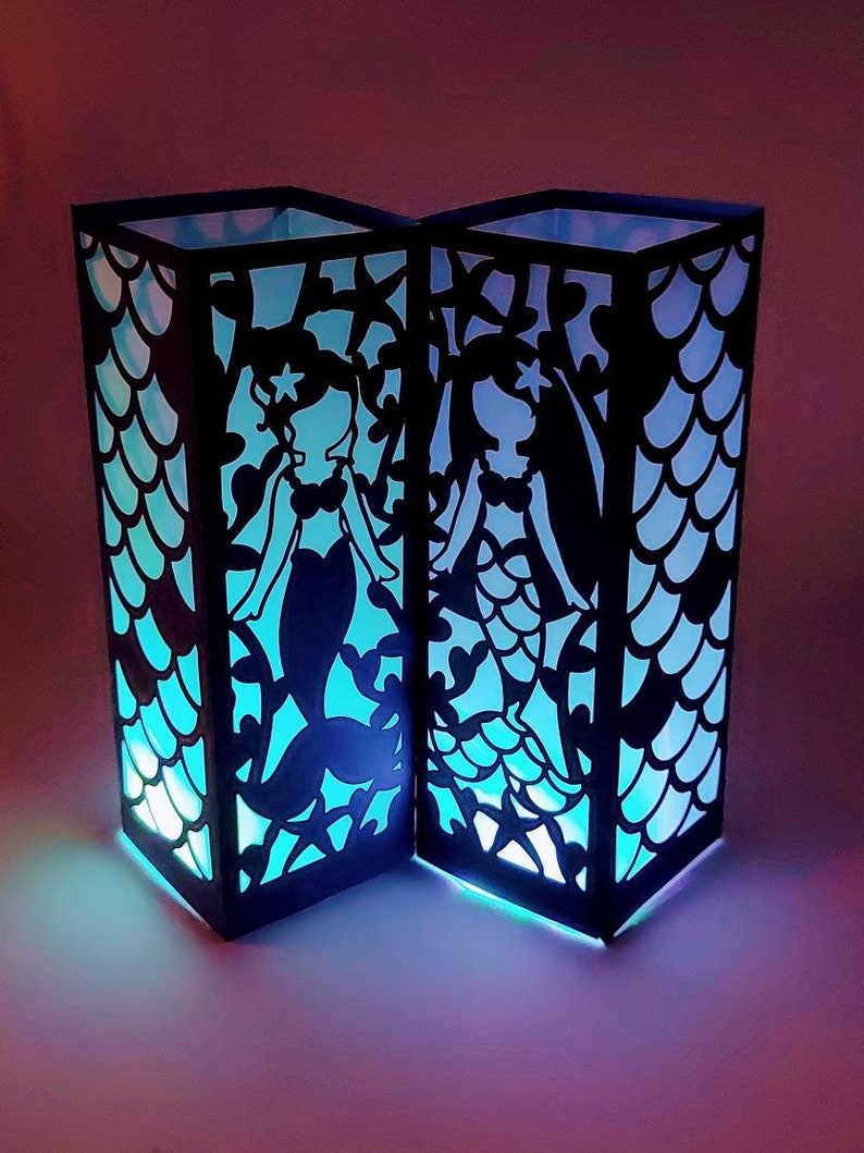 Puede incluir: Dos linternas de papel negro con siluetas de sirenas y un patr&oacute;n de escamas. Las linternas est&aacute;n iluminadas desde el interior, proyectando un brillo azul y morado.