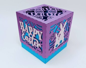 May include: Purple and blue papercraft box with a cut-out design of bunnies and eggs. The box has the words "Happy Easter" on the front.