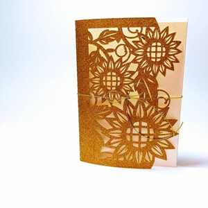 May include: Gold glitter laser cut card with a sunflower design. The card is folded in half and tied with a gold ribbon.