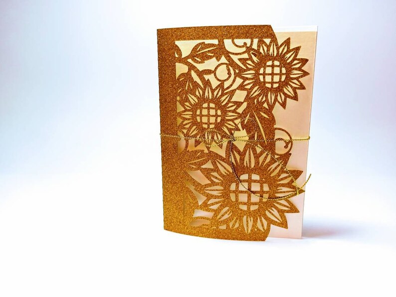 May include: Gold glitter laser cut card with a sunflower design. The card is folded in half and tied with a gold ribbon.