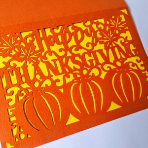 Puede incluir: Tarjeta naranja con fondo amarillo y un dise&ntilde;o recortado de calabazas y las palabras "Feliz D&iacute;a de Acci&oacute;n de Gracias".