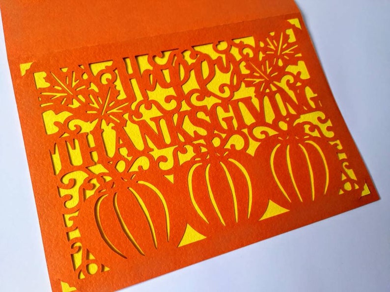 Puede incluir: Tarjeta naranja con fondo amarillo y un dise&ntilde;o recortado de calabazas y las palabras "Feliz D&iacute;a de Acci&oacute;n de Gracias".