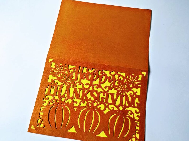 Puede incluir: Tarjeta naranja con un inserto amarillo. La tarjeta tiene un dise&ntilde;o recortado de calabazas, hojas y las palabras "Feliz D&iacute;a de Acci&oacute;n de Gracias".