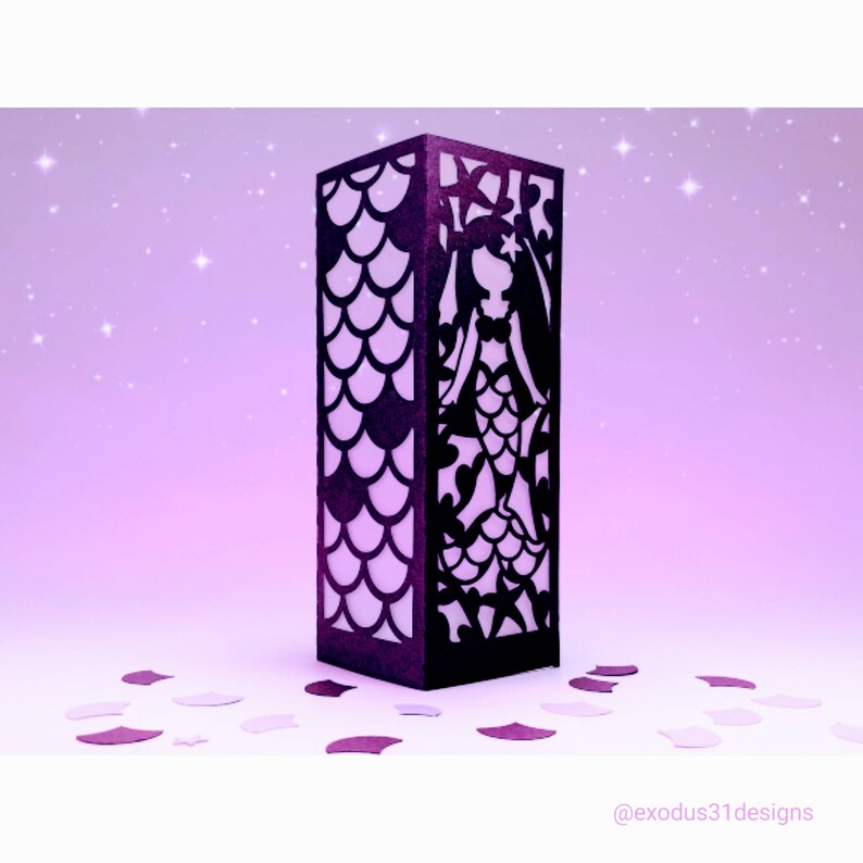 Puede incluir: Una linterna de papel negra con una silueta de sirena y un patr&oacute;n de escamas. La linterna est&aacute; sobre un fondo morado con purpurina.