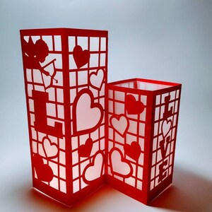 May include: Two red paper lanterns with cut-out designs of hearts and the word "LOVE". The lanterns are illuminated from within, creating a warm glow.