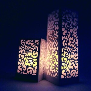 May include: Three white paper lanterns with intricate cut-out designs. The lanterns are lit from within, casting a warm glow.