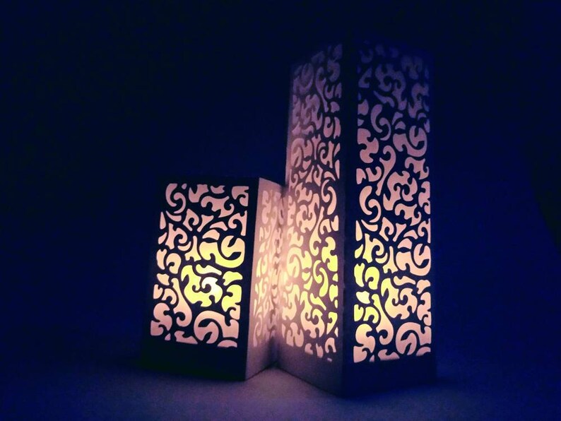 May include: Three white paper lanterns with intricate cut-out designs. The lanterns are lit from within, casting a warm glow.