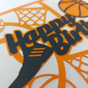 May include: A white card with an orange and black basketball theme. The card has a black cutout of the words "Happy Birthday" with a basketball and net design in the background.