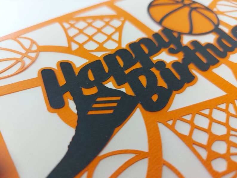 May include: A white card with an orange and black basketball theme. The card has a black cutout of the words "Happy Birthday" with a basketball and net design in the background.