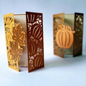 May include: Two brown paper cards with intricate cut-out designs of pumpkins and leaves. The cards are folded and standing upright on a white surface.