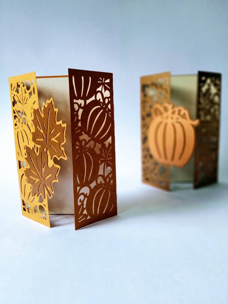 May include: Two brown paper cards with intricate cut-out designs of pumpkins and leaves. The cards are folded and standing upright on a white surface.
