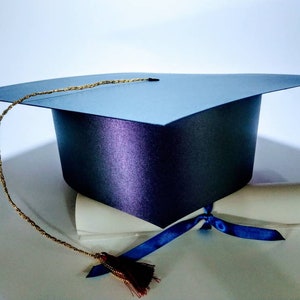Puede incluir: Un gorro de graduaci&oacute;n negro con una borla dorada y un diploma blanco atado con una cinta azul.