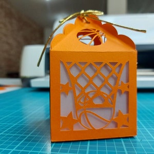 May include: Orange paper gift box with a cut-out design of a basketball hoop and basketball. The box has a gold ribbon.