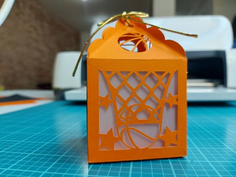 May include: Orange paper gift box with a cut-out design of a basketball hoop and basketball. The box has a gold ribbon.