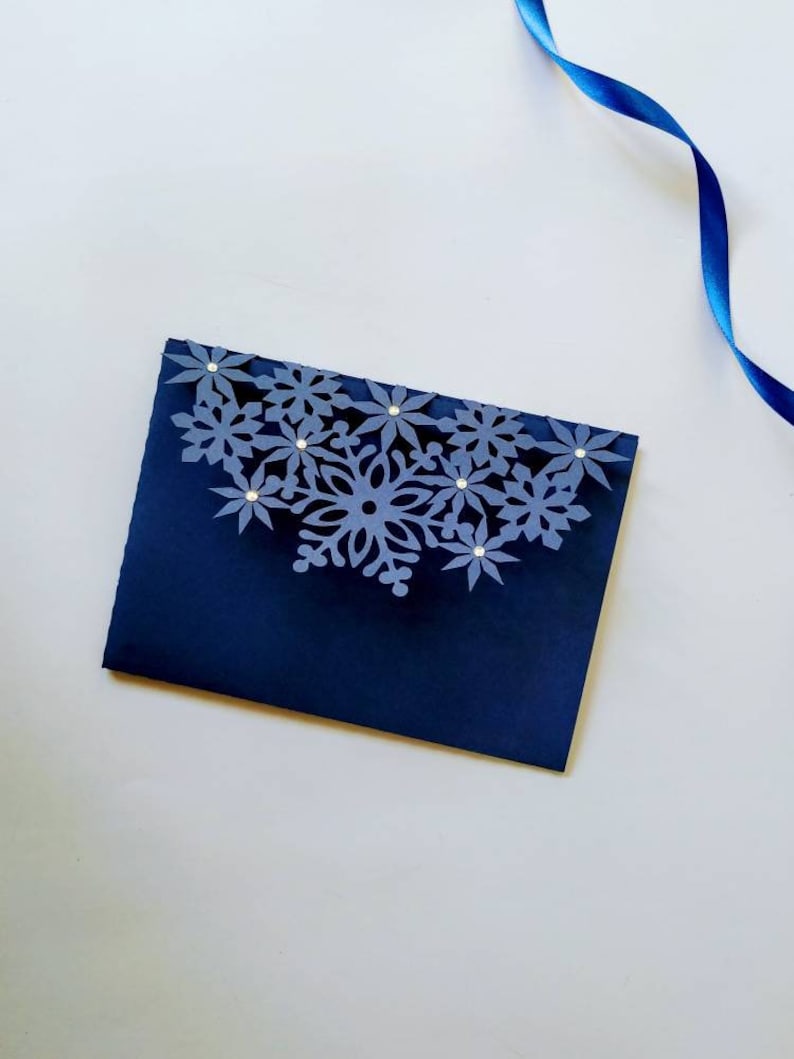 K&ouml;nnte beinhalten: Ein dunkelblauer Umschlag mit einem wei&szlig;en Schneeflockenmuster. Die Schneeflocke ist aus Papier und hat einen 3D-Effekt. Der Umschlag ist mit kleinen, klaren Strasssteinen verziert.