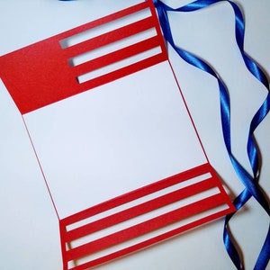 May include: A red card with white stripes cut out, folded in half, with a blue ribbon on a white background.