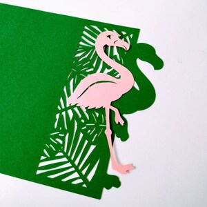 May include: A pink flamingo cutout with a green tropical leaf background. The flamingo is standing on one leg and has its head turned to the side. The leaves are cut out of the green paper, creating a layered effect.