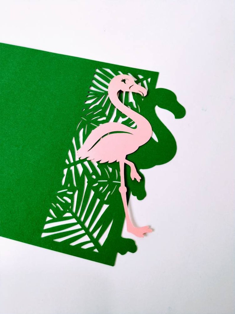 May include: A pink flamingo cutout with a green tropical leaf background. The flamingo is standing on one leg and has its head turned to the side. The leaves are cut out of the green paper, creating a layered effect.