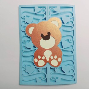May include: A light blue card with a cut-out design featuring a brown teddy bear with white paws. The card is decorated with hearts and swirls.