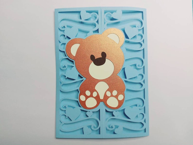 May include: A light blue card with a cut-out design featuring a brown teddy bear with white paws. The card is decorated with hearts and swirls.