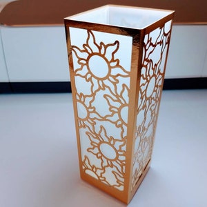 May include: A white paper lantern with a gold metallic border and a cut-out design of suns with rays. The lantern is sitting on a white surface.