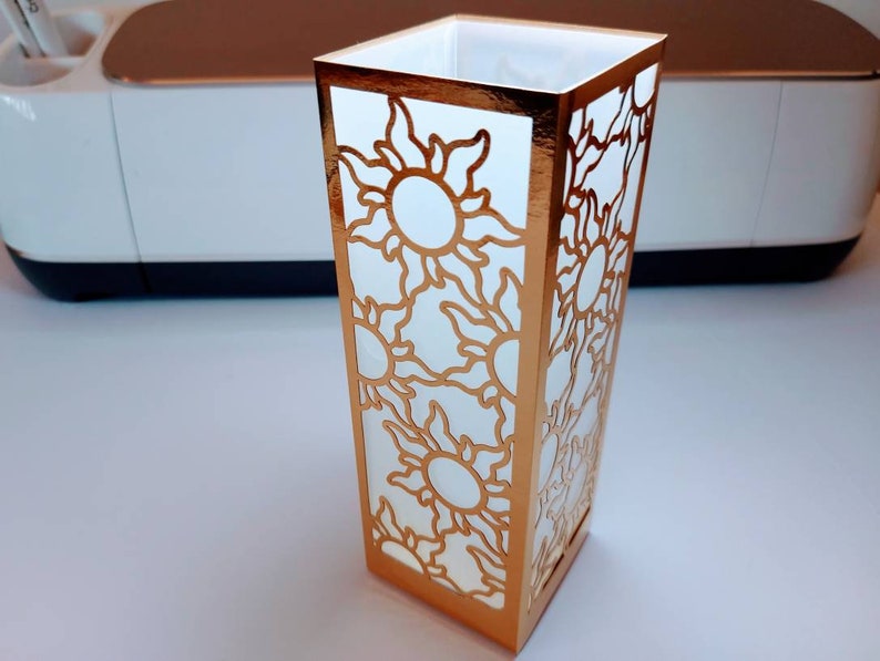 May include: A white paper lantern with a gold metallic border and a cut-out design of suns with rays. The lantern is sitting on a white surface.