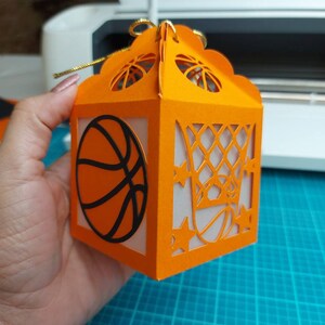 May include: Orange paper gift box with a basketball and basketball hoop design. The box has a gold ribbon.