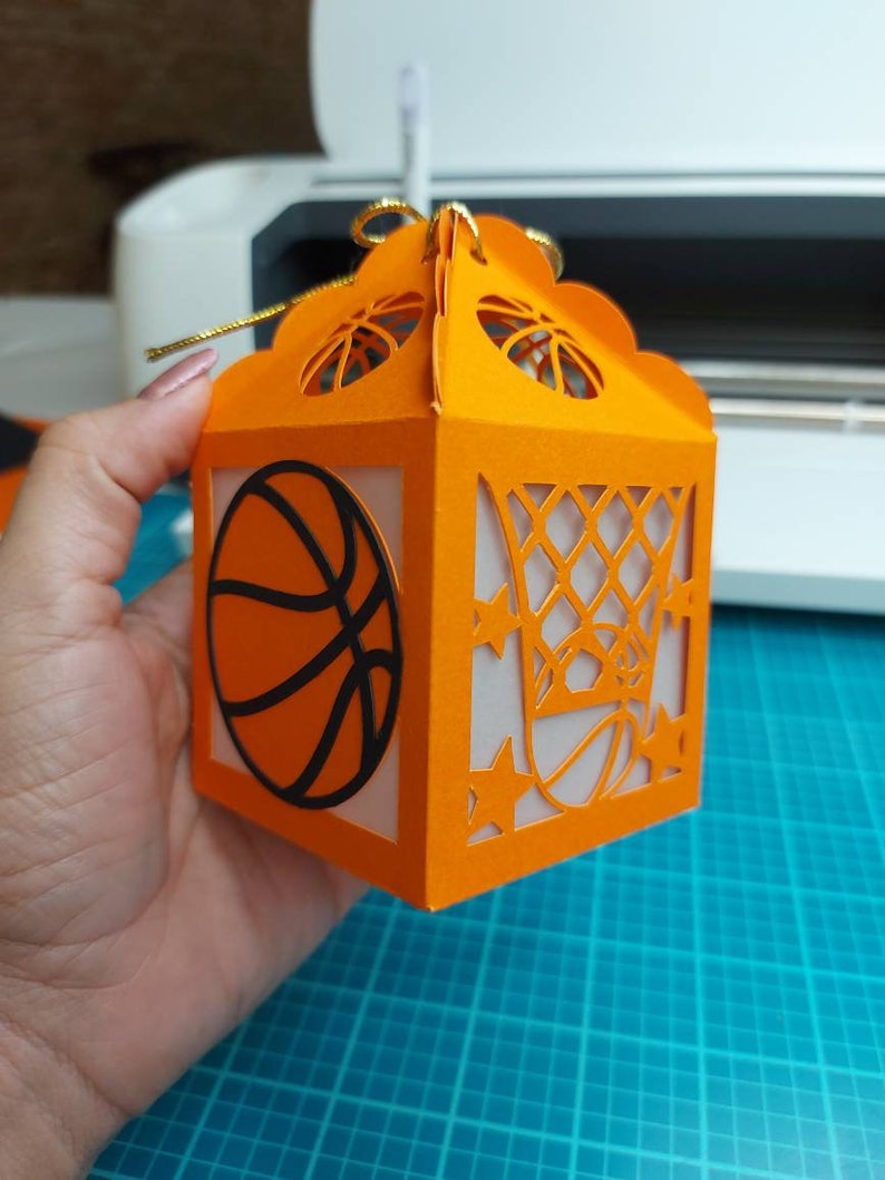 May include: Orange paper gift box with a basketball and basketball hoop design. The box has a gold ribbon.