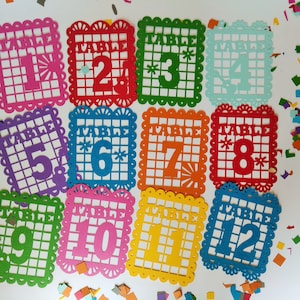 May include: Twelve colorful paper table numbers, each with a different color and number from 1 to 12. The numbers are in a square shape with a decorative border. The numbers are perfect for a party or event.