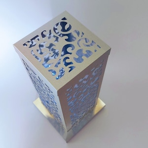 Puede incluir: Una linterna rectangular dorada con intrincados diseños calados. La linterna tiene una base y una parte superior cuadradas, con un interior hueco. Los patrones calados crean un efecto decorativo, y la linterna parece ser de metal.