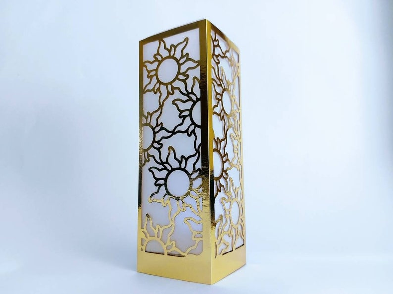 May include: A gold-colored paper lantern with a cut-out design of multiple suns. The lantern is made of paper and has a white interior.
