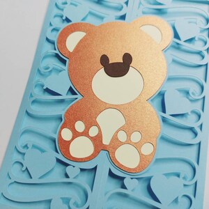 May include: A light blue card with a decorative cut-out design featuring hearts and swirls. A copper-colored teddy bear with white paws is centered on the card.