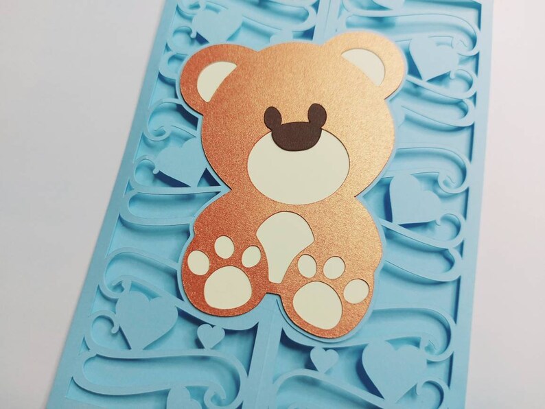 May include: A light blue card with a decorative cut-out design featuring hearts and swirls. A copper-colored teddy bear with white paws is centered on the card.