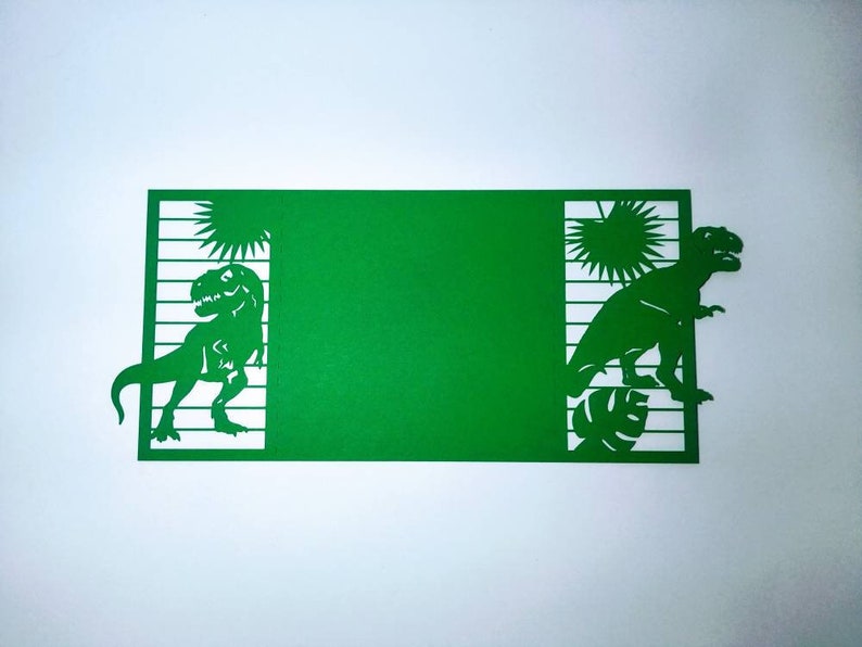 May include: Green cardstock cut-out with two dinosaurs, one on each side. The dinosaurs are facing each other and are surrounded by palm trees and leaves. The cardstock is blank in the middle.