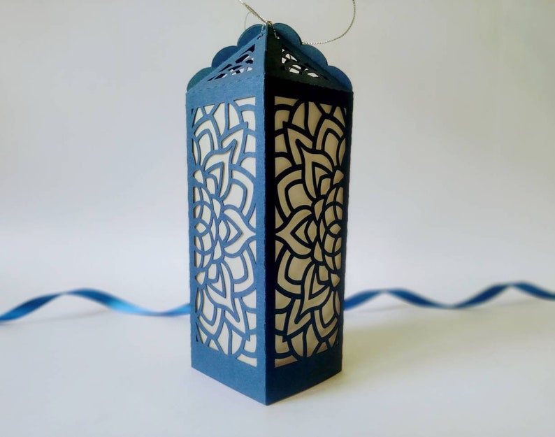 May include: A blue paper lantern with a white intricate design cut out. The lantern is hanging from a string.