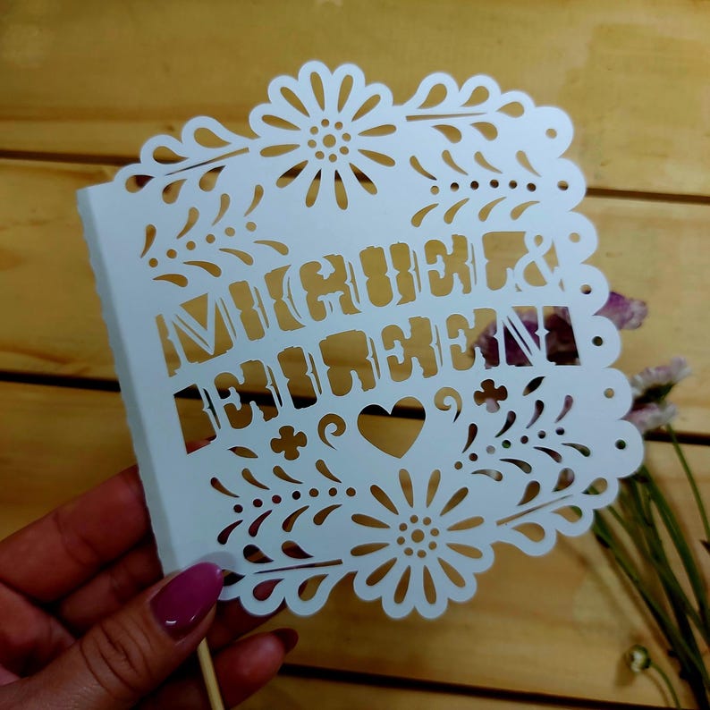 May include: A white paper cake topper with the names "Miguel & Lorena" cut out in a decorative font. The topper features floral and heart cutouts along the edges and is attached to a wooden stick.