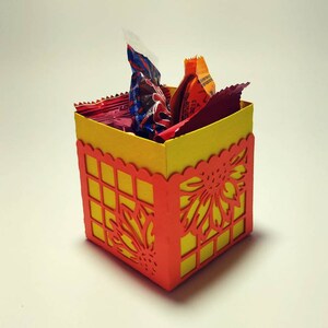 Puede incluir: Una peque&ntilde;a caja de papel amarilla y naranja con un dise&ntilde;o decorativo de corte. La caja est&aacute; llena de caramelos envueltos.