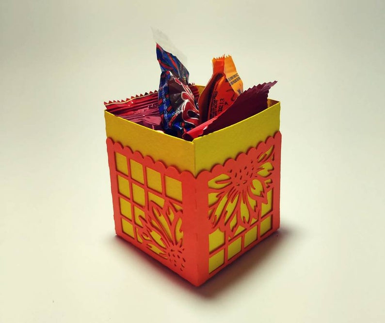 Puede incluir: Una peque&ntilde;a caja de papel amarilla y naranja con un dise&ntilde;o decorativo de corte. La caja est&aacute; llena de caramelos envueltos.