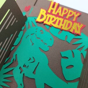 May include: A green dinosaur silhouette with a brown background and the words "Happy Birthday" in red and yellow letters.