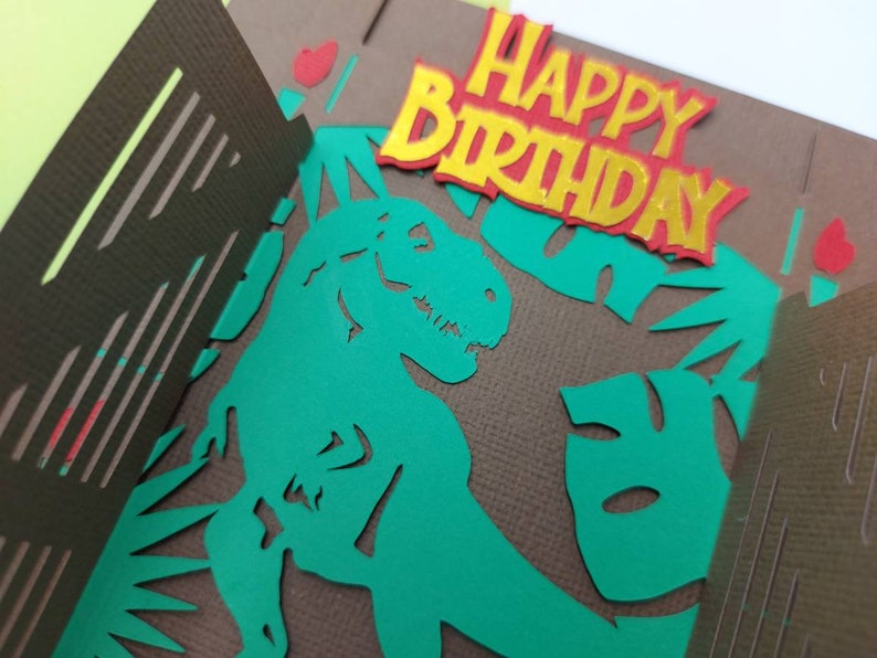 May include: A green dinosaur silhouette with a brown background and the words "Happy Birthday" in red and yellow letters.