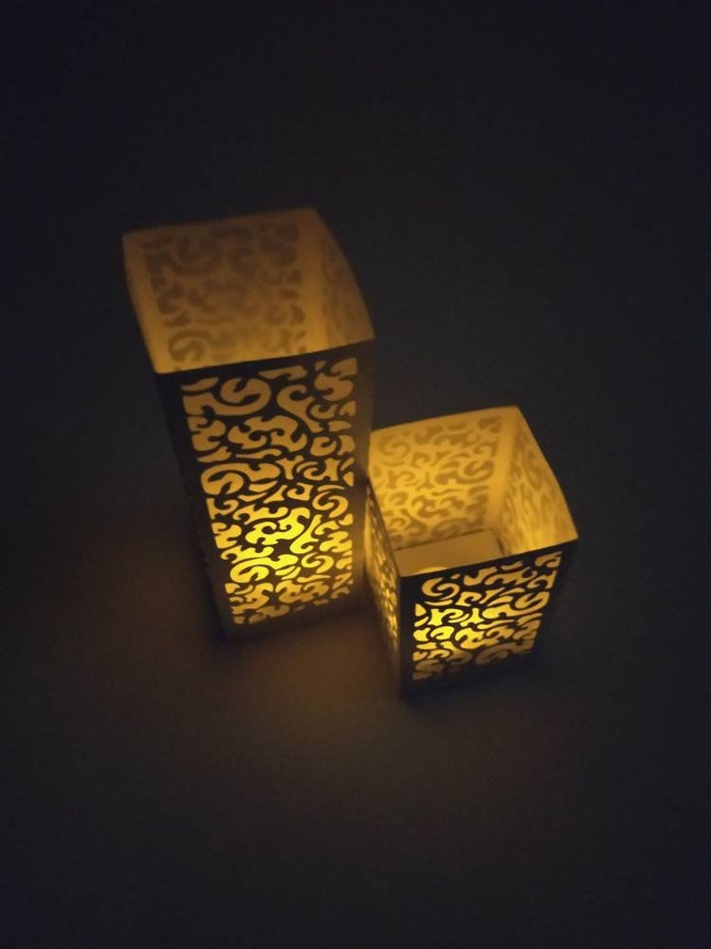 May include: Two white paper candle holders with a decorative cut-out design. The candle holders are lit and casting a warm glow.