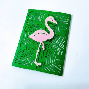 May include: A green card with a pink flamingo cutout. The card is decorated with a laser cut design of tropical leaves.